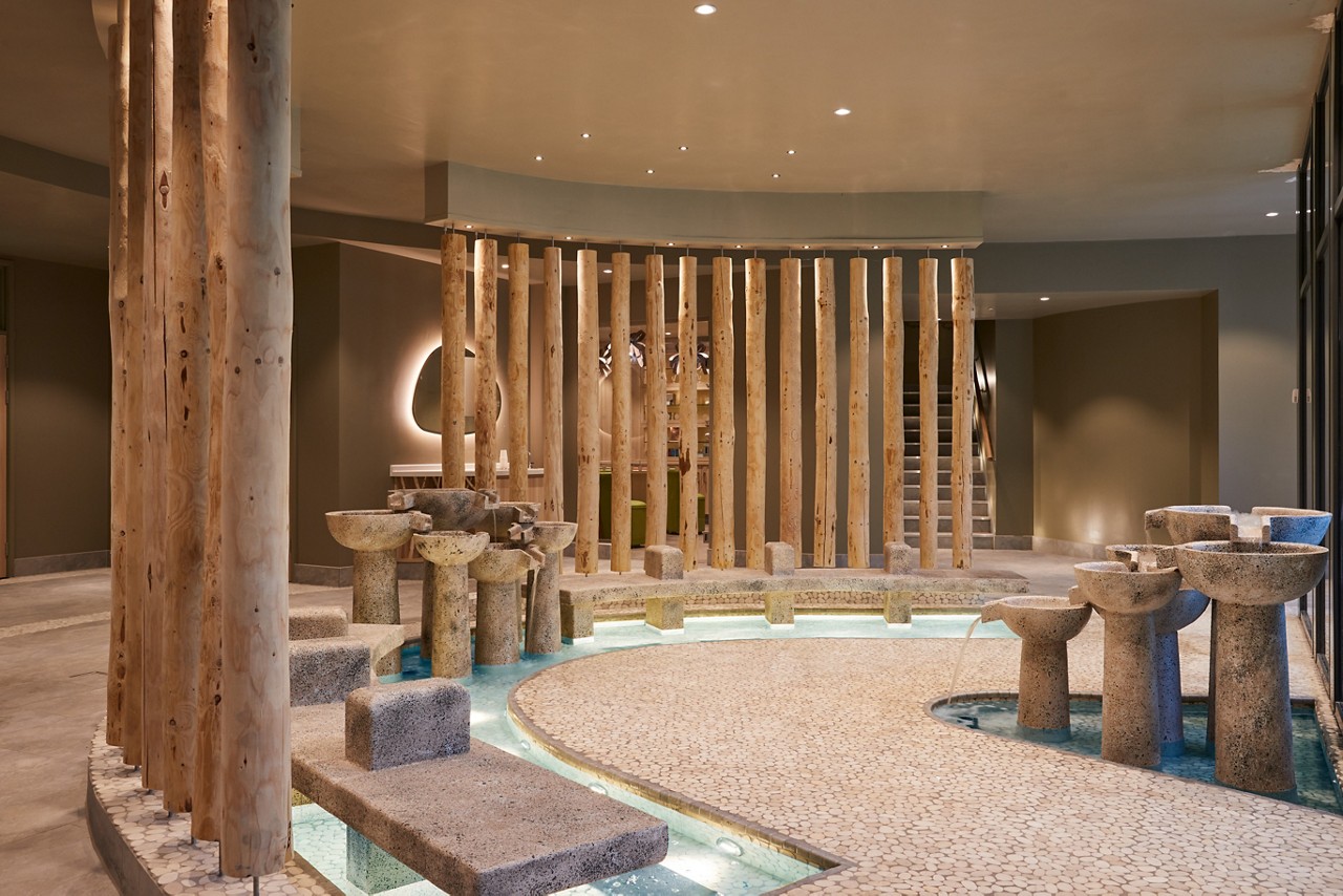 Stone basins trickle water into shallow channels within a curved, pebble-floored spa area. Wooden columns partition the space, while warm recessed lighting and nearby stairs create a serene, modern interior.